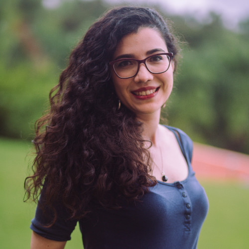 Foto de perfil Gabriela Feijó. Gabriela está sorrindo e vestindo uma blusa azul. O fundo da foto está desfocado, mas parece um lugar com árvores